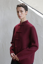 Load image into Gallery viewer, A woman with short dark hair wears the Mandarin Jacket | Watermark Burgundy, featuring toggle closures, standing against a light-textured wall and looking calmly at the camera.
