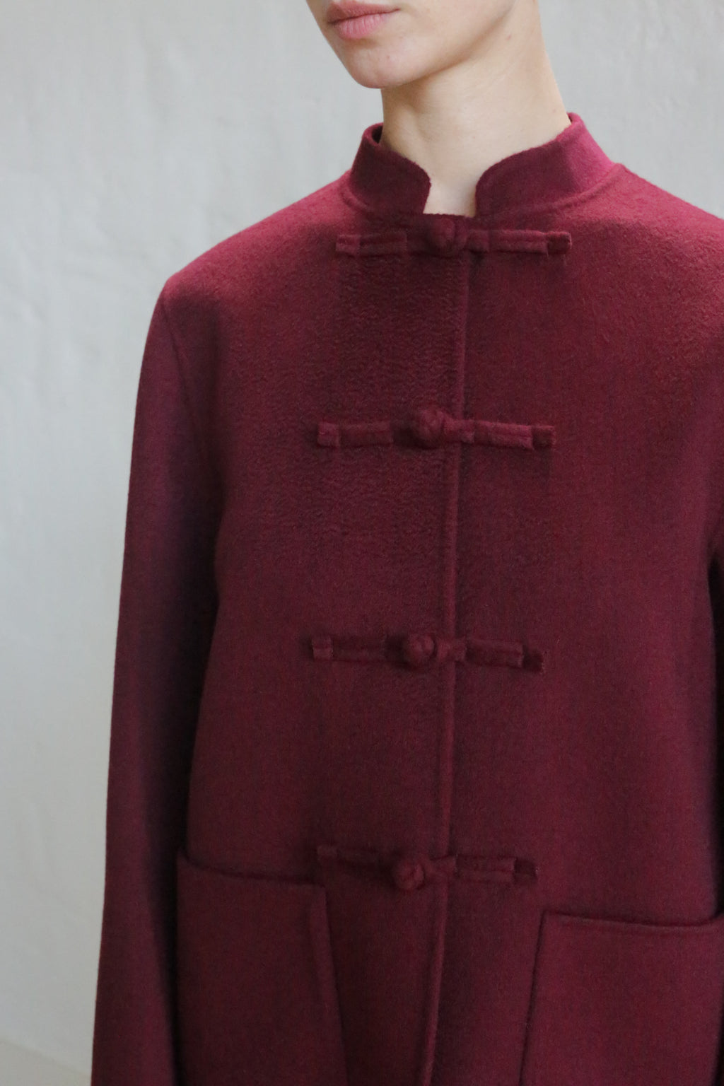 Someone wears the Mandarin Jacket | Watermark Burgundy, a maroon, high-collared coat with traditional frog buttons, shown from shoulders to mid-torso against a plain light background.