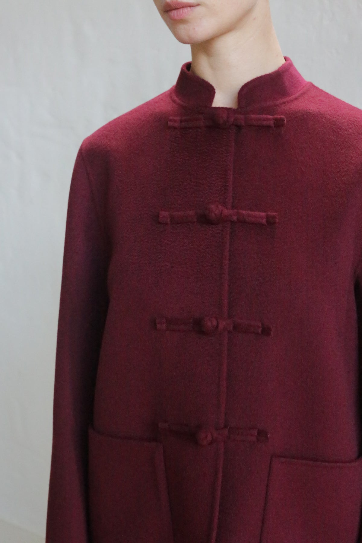 Someone wears the Mandarin Jacket | Watermark Burgundy, a maroon, high-collared coat with traditional frog buttons, shown from shoulders to mid-torso against a plain light background.