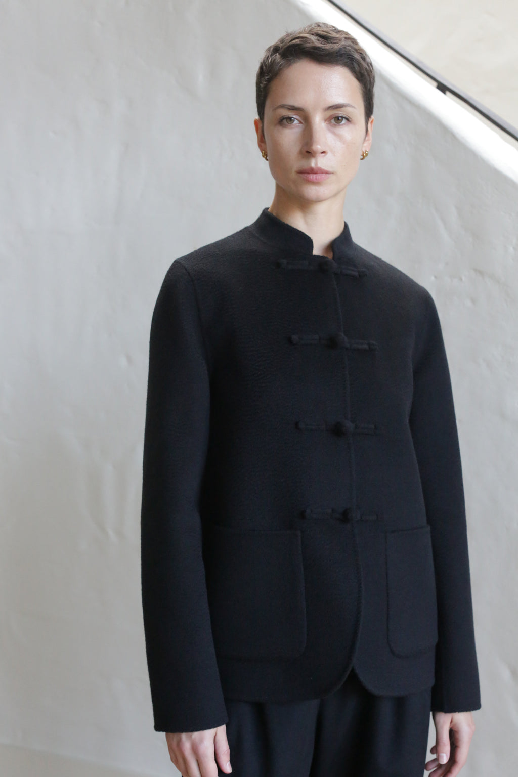 A person with short dark hair wears the Mandarin Jacket | Watermark Black, a collarless, buttoned jacket with two front pockets, standing against a light-colored textured wall.