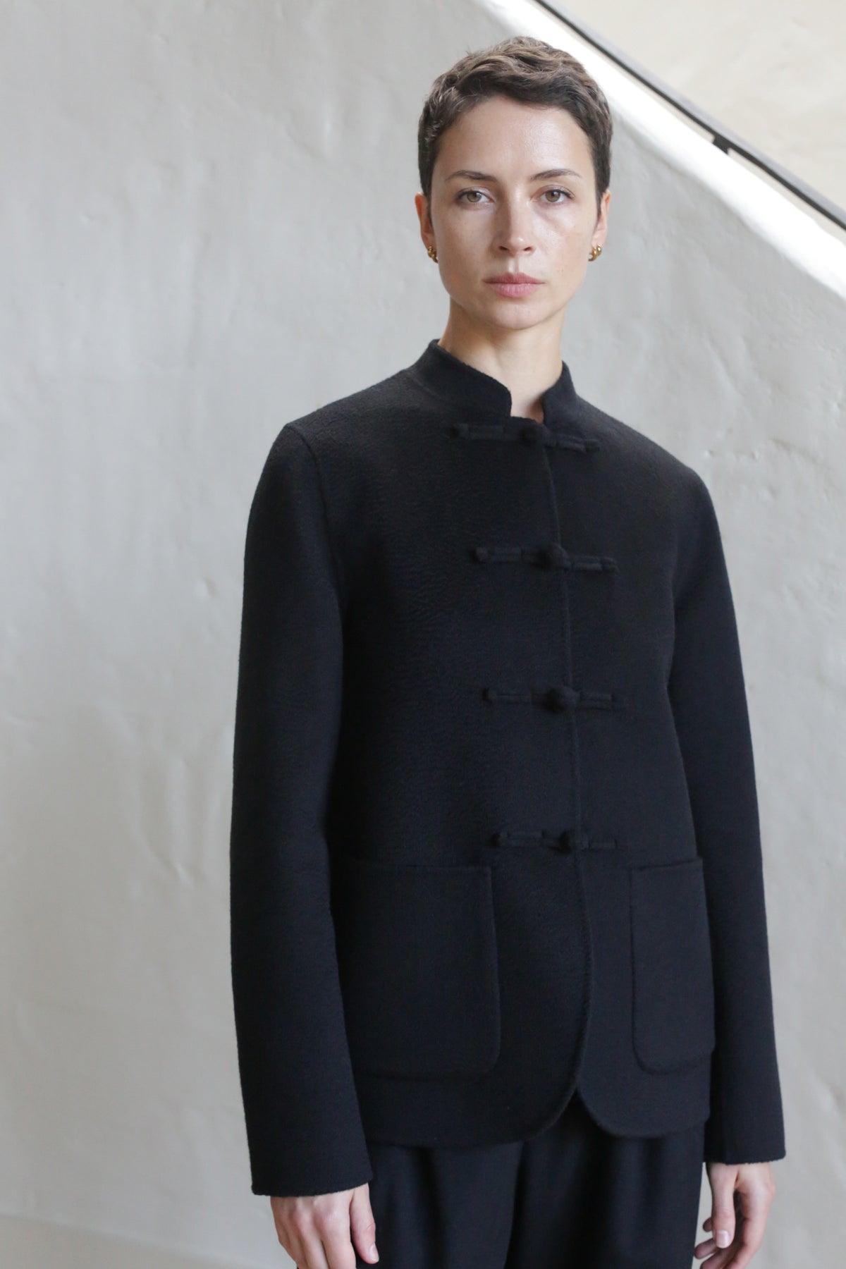 A person with short dark hair wears the Mandarin Jacket | Watermark Black, a collarless, buttoned jacket with two front pockets, standing against a light-colored textured wall.