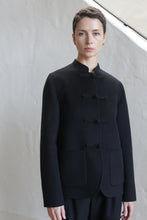 Load image into Gallery viewer, A person with short dark hair wears the Mandarin Jacket | Watermark Black, a collarless, buttoned jacket with two front pockets, standing against a light-colored textured wall.