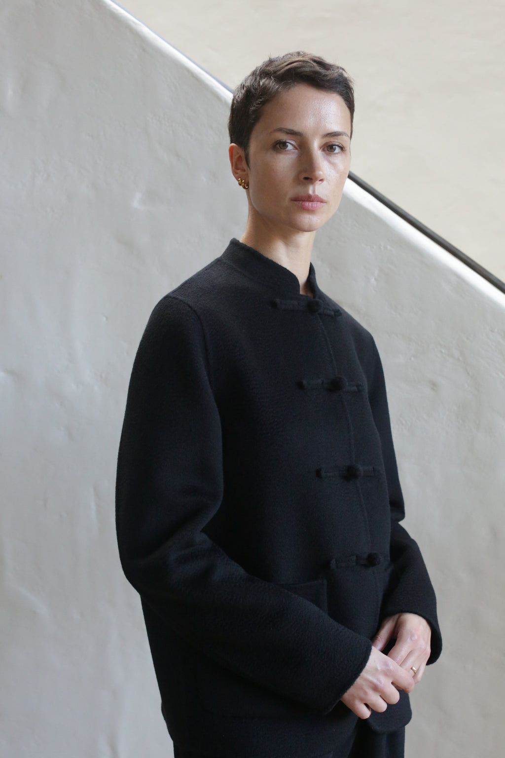 A person with short dark hair wears the Mandarin Jacket | Watermark Black, standing indoors against a light-colored textured wall and looking calmly at the camera.