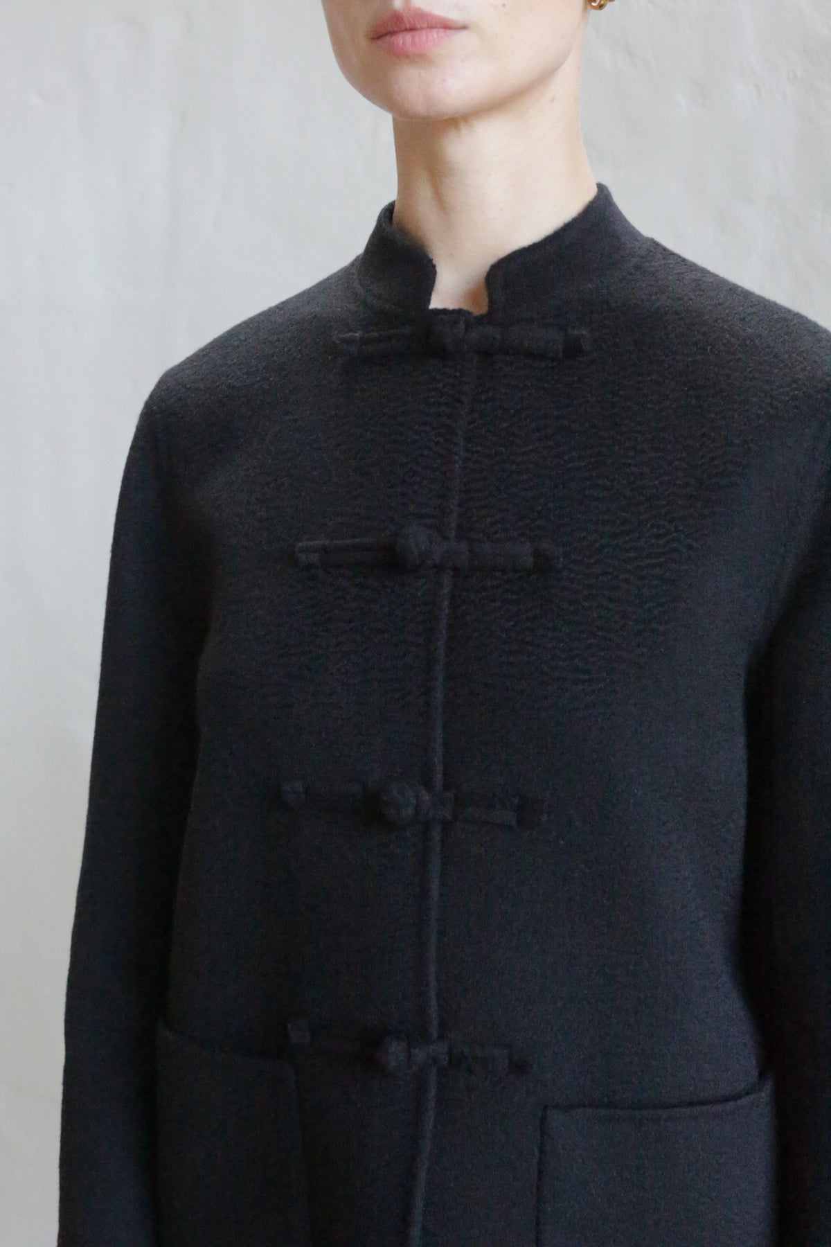 A person models the Mandarin Jacket | Watermark Black, featuring a black mandarin collar, traditional knotted buttons, and two front pockets, shown from shoulders to waist against a plain light background.