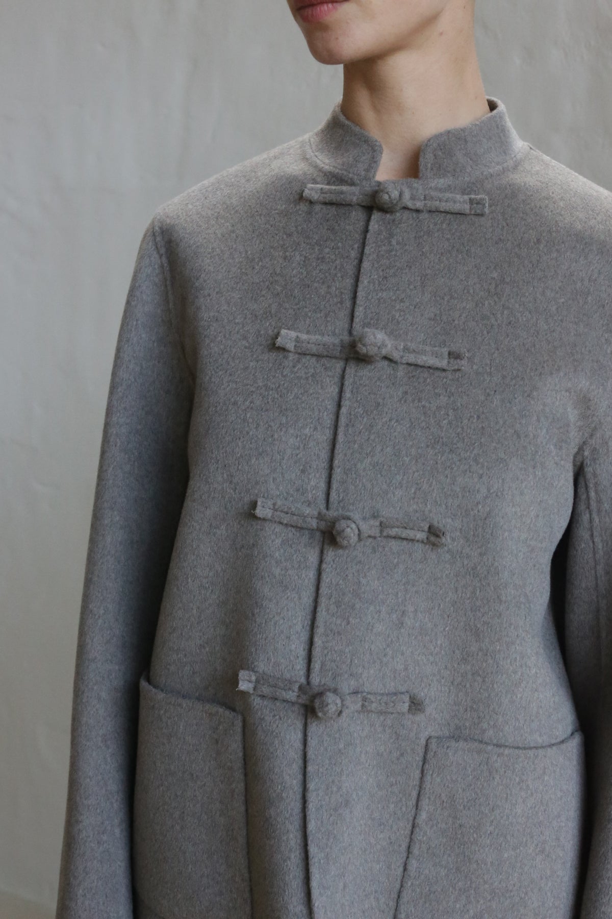 A person models the Mandarin Jacket | Undyed Natural, featuring a high collar, three frog button closures, and two large front pockets, set against a plain light background.