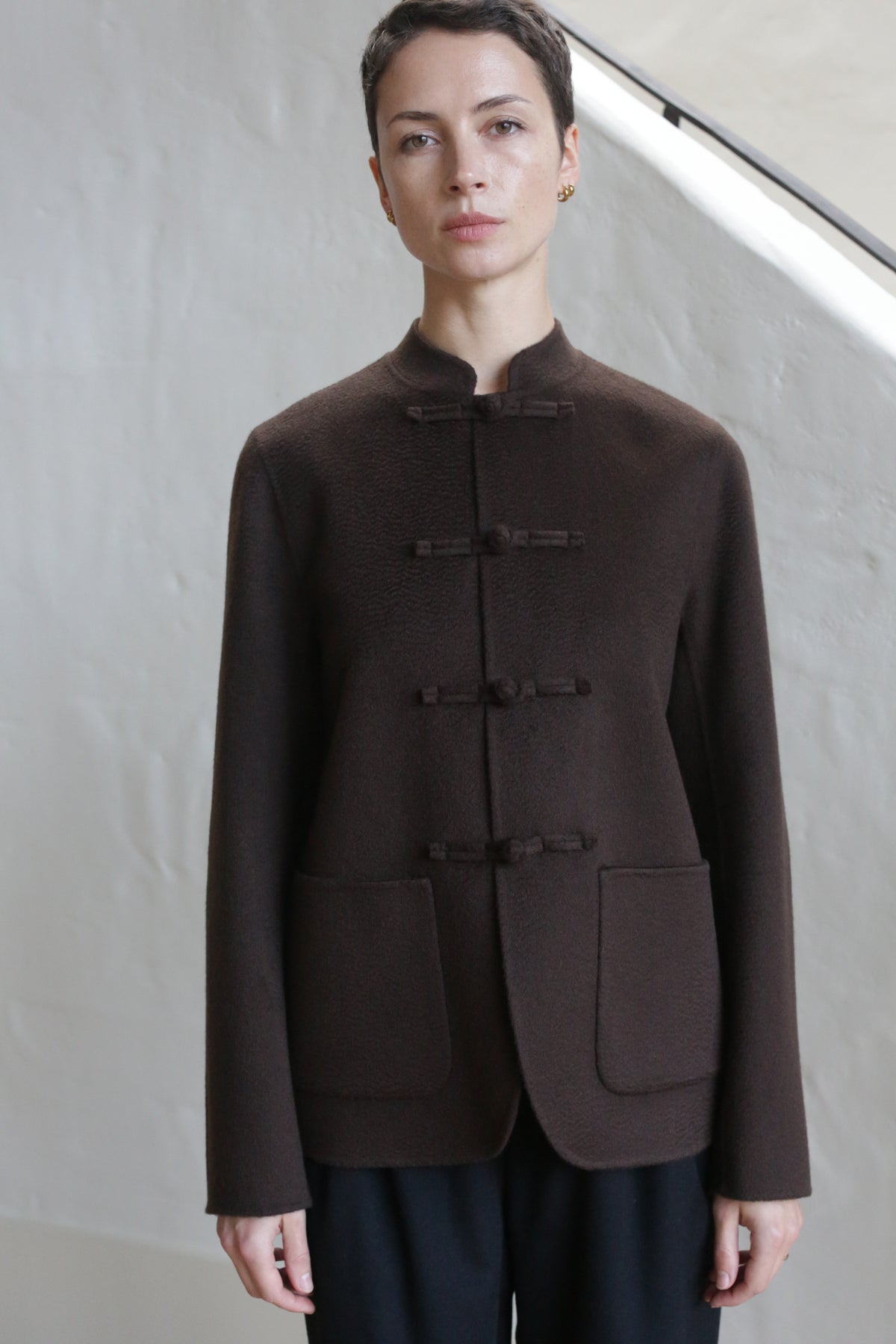 A person with short hair wears the Mandarin Jacket in Watermark Brown, featuring a high neck, cloth toggle closures, and large front pockets, standing in front of a light-colored wall.