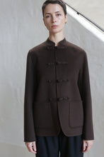 Load image into Gallery viewer, A person with short hair wears the Mandarin Jacket in Watermark Brown, featuring a high neck, cloth toggle closures, and large front pockets, standing in front of a light-colored wall.