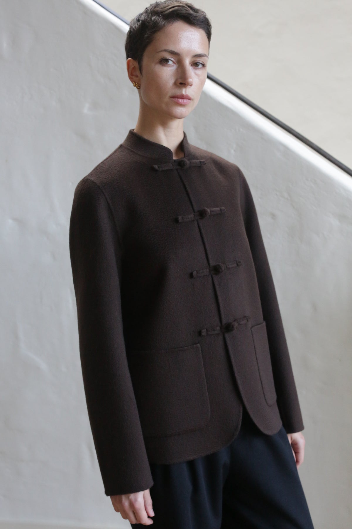 A person with short hair wears the Mandarin Jacket | Watermark Brown, buttoned up, paired with black pants, standing before a light textured wall with a diagonal railing in the background.