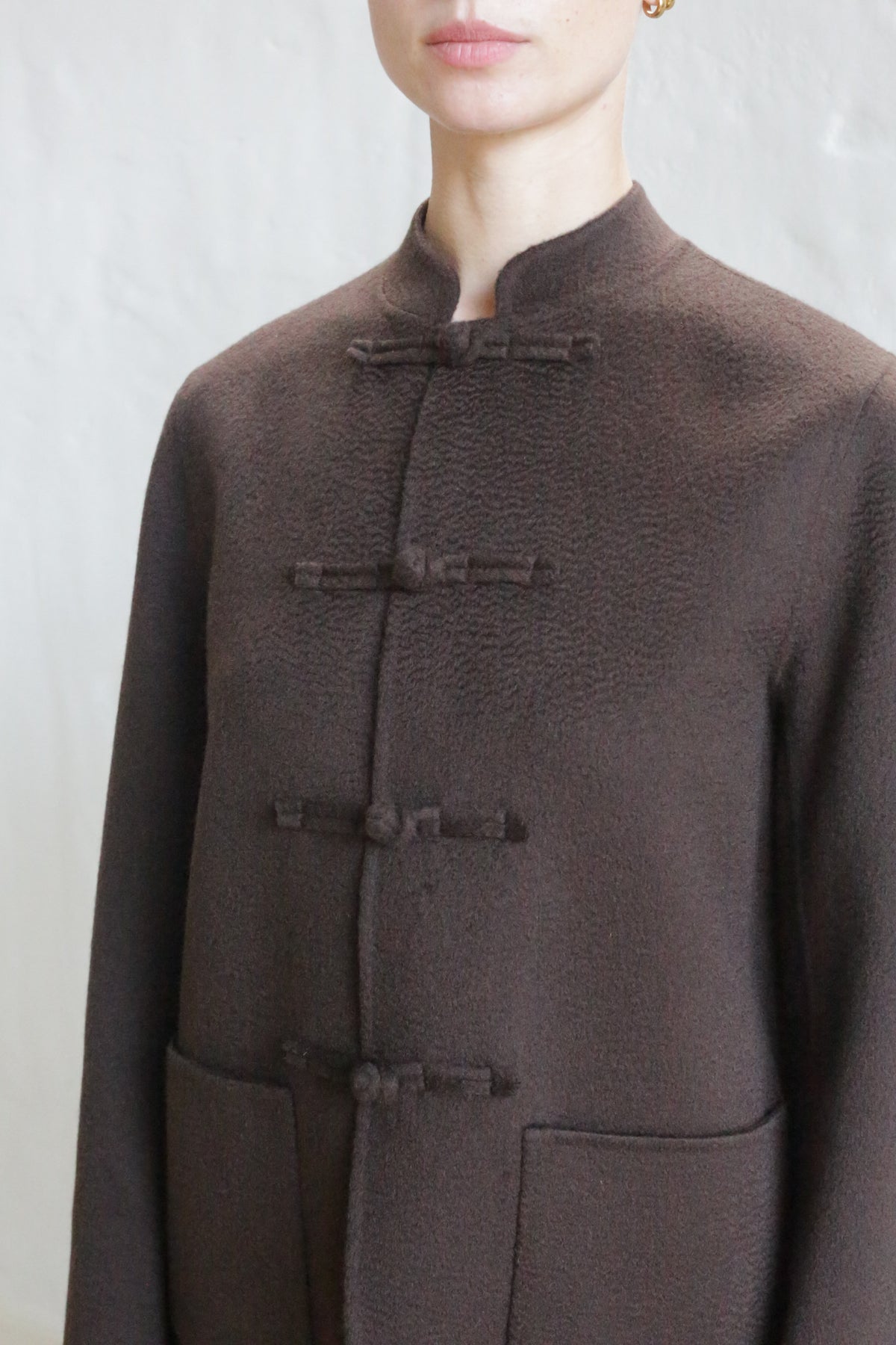 A person models the Mandarin Jacket | Watermark Brown, featuring a textured brown fabric, Mandarin collar, and three knotted toggle closures, shown from shoulders to waist against a light background.
