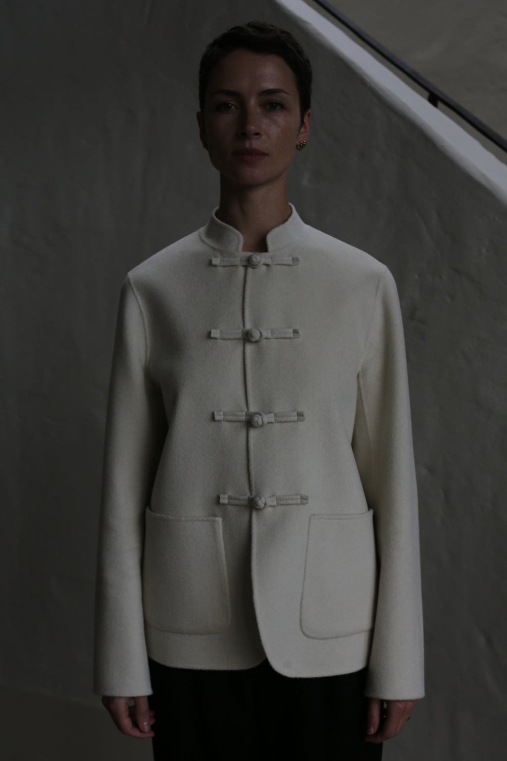 A person stands against a gray wall, wearing the Mandarin Jacket | Undyed—a white, long-sleeved jacket with a mandarin collar and knotted buttons. Dim lighting casts shadows on their face and upper body.
