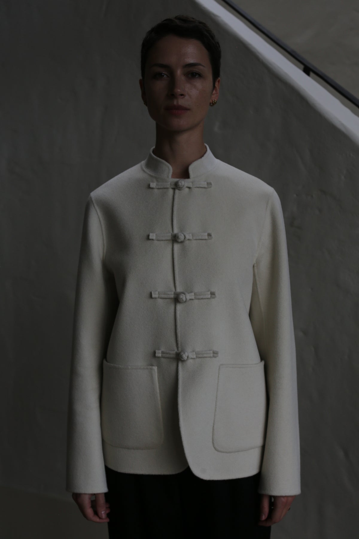 A person stands against a gray wall, wearing the Mandarin Jacket | Undyed—a white, long-sleeved jacket with a mandarin collar and knotted buttons. Dim lighting casts shadows on their face and upper body.
