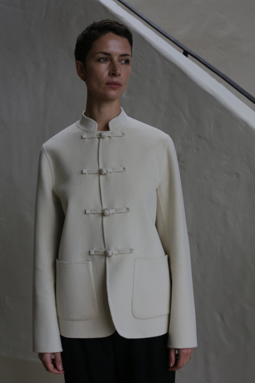 A person with short dark hair wears the Mandarin Jacket | Undyed, featuring a cream hue and large front pockets, while standing before a neutral textured wall and staircase railing.