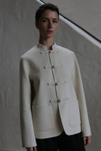 Load image into Gallery viewer, A person with short dark hair wears the Mandarin Jacket | Undyed, a cream-colored, collarless jacket with four toggle closures and large front pockets, standing against a textured, neutral wall.