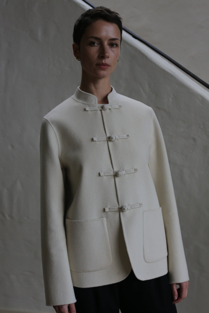 A person with short dark hair wears the Mandarin Jacket | Undyed, a cream-colored, collarless jacket with four toggle closures and large front pockets, standing against a textured, neutral wall.