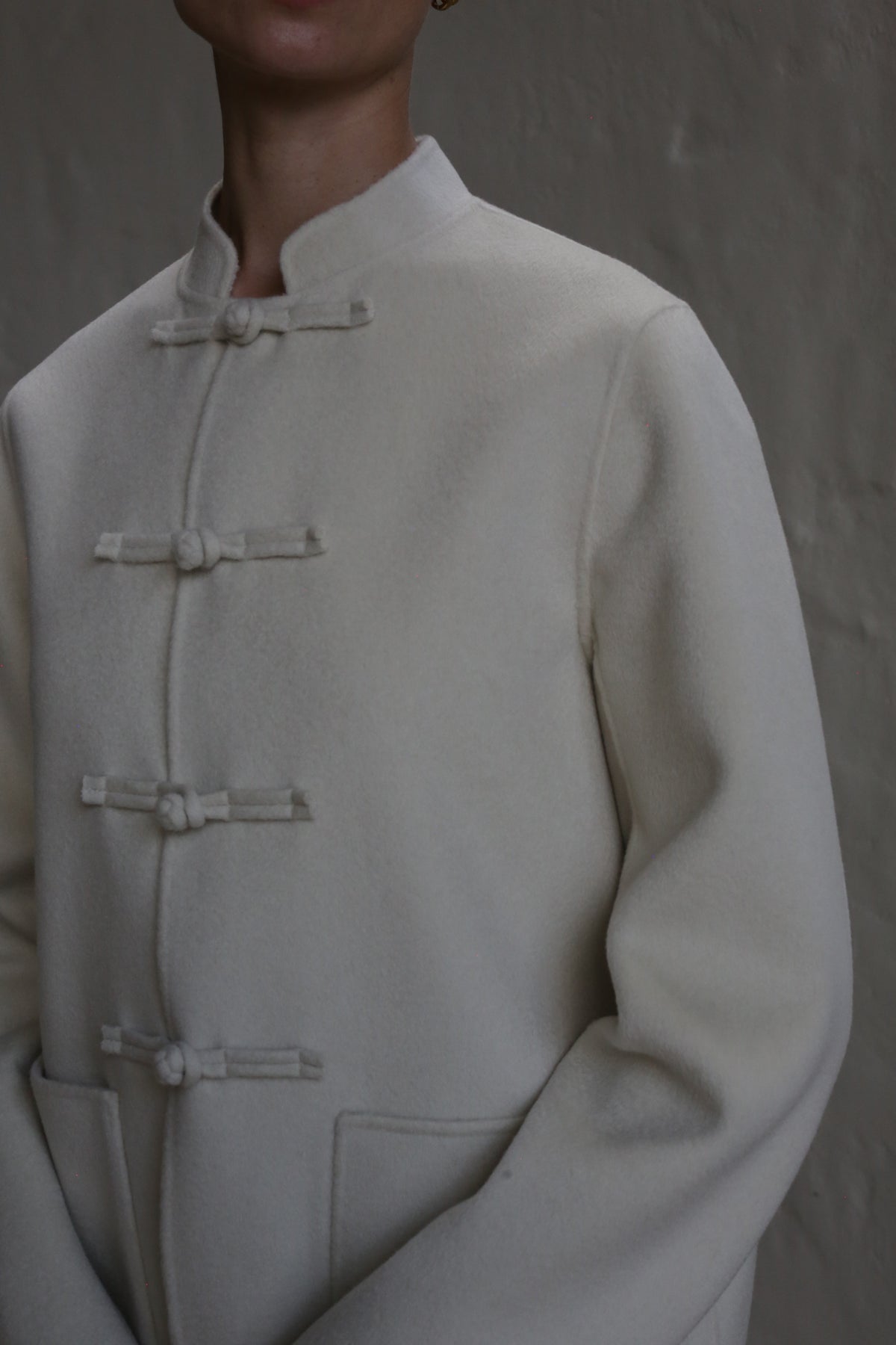 A person models the Mandarin Jacket | Undyed, a cream-colored, high-collared coat with three knotted button closures, standing against a plain textured background. The image is cropped at the neck and waist.
