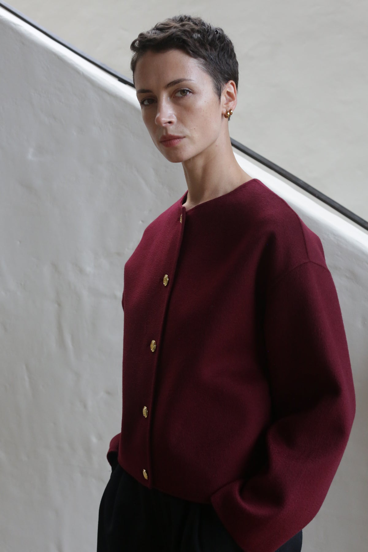 A person with short dark hair wears the Cocoon Jacket in Watermark Burgundy and gold earrings, standing in front of a textured white wall with a black railing behind them.