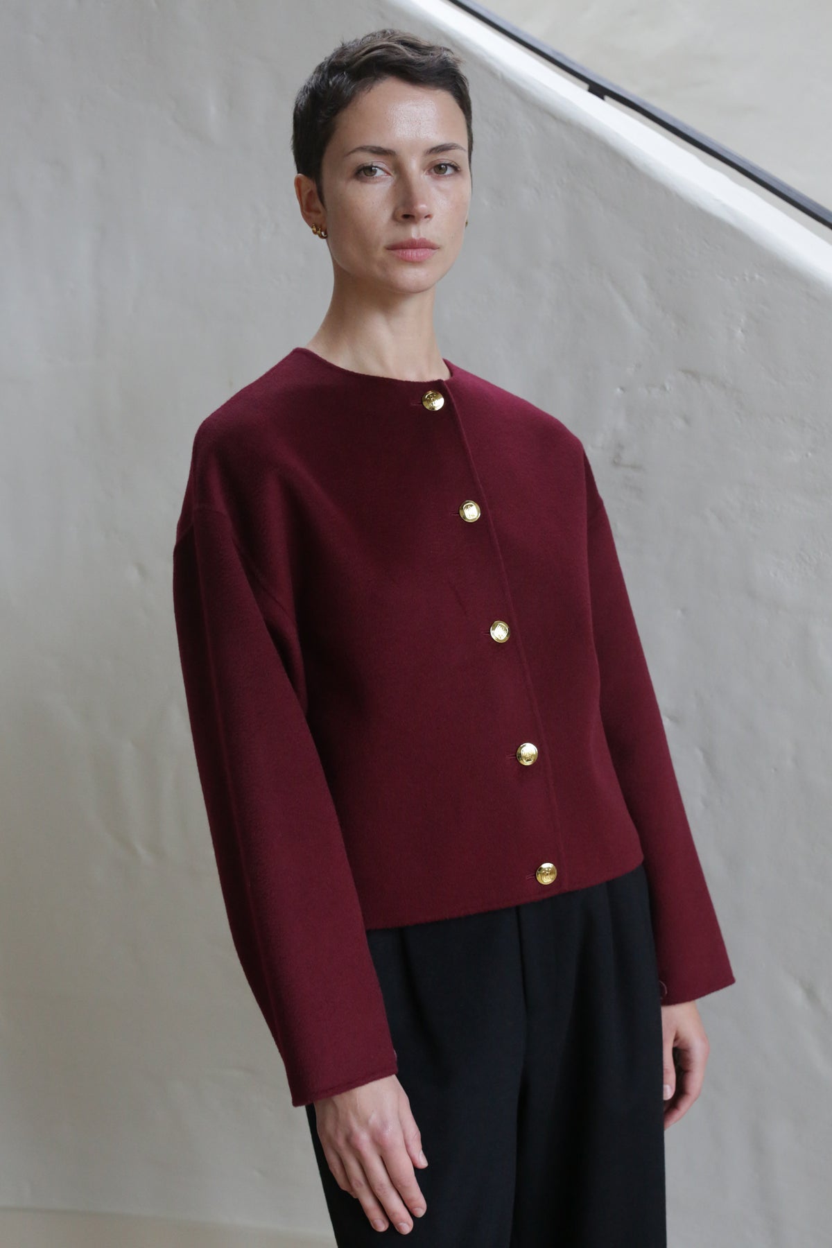 A person with short dark hair wears the Cocoon Jacket | Watermark Burgundy and black pants, standing by a light textured wall near a staircase banister.