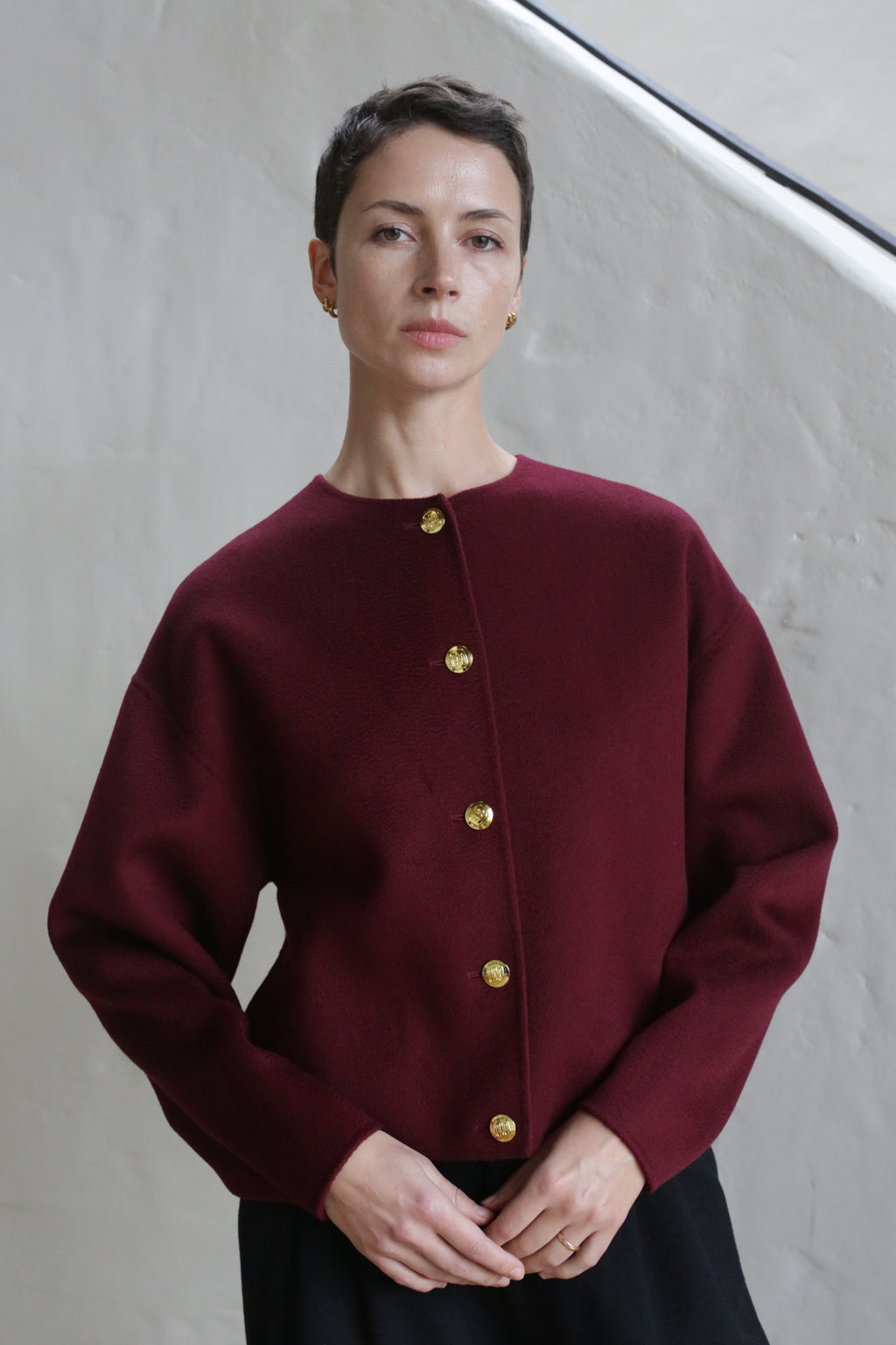 A person with short brown hair wears the Cocoon Jacket | Watermark Burgundy, featuring gold buttons, and stands calmly before a light-colored textured wall.