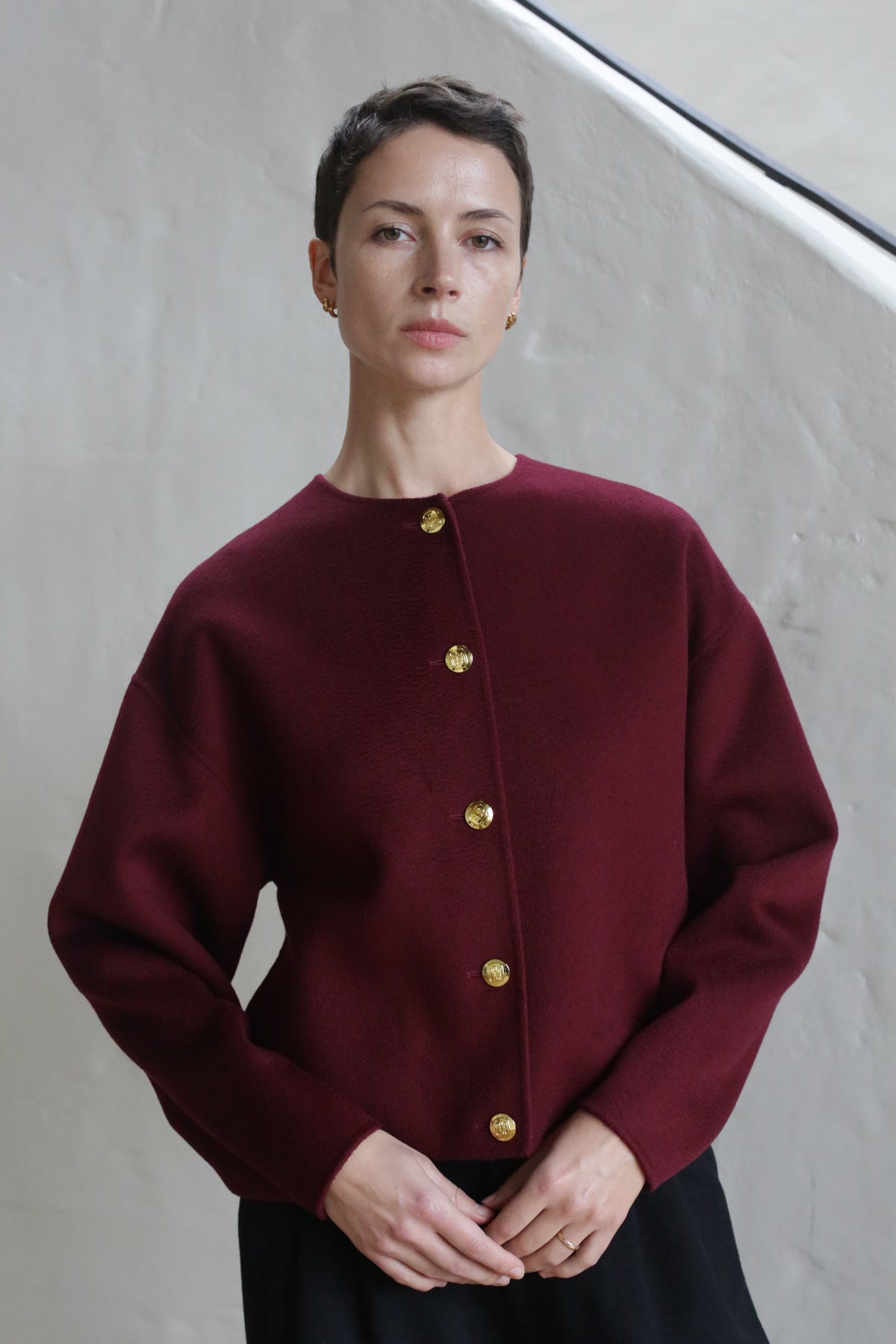 A person with short brown hair wears the Cocoon Jacket | Watermark Burgundy, featuring gold buttons, and stands calmly before a light-colored textured wall.