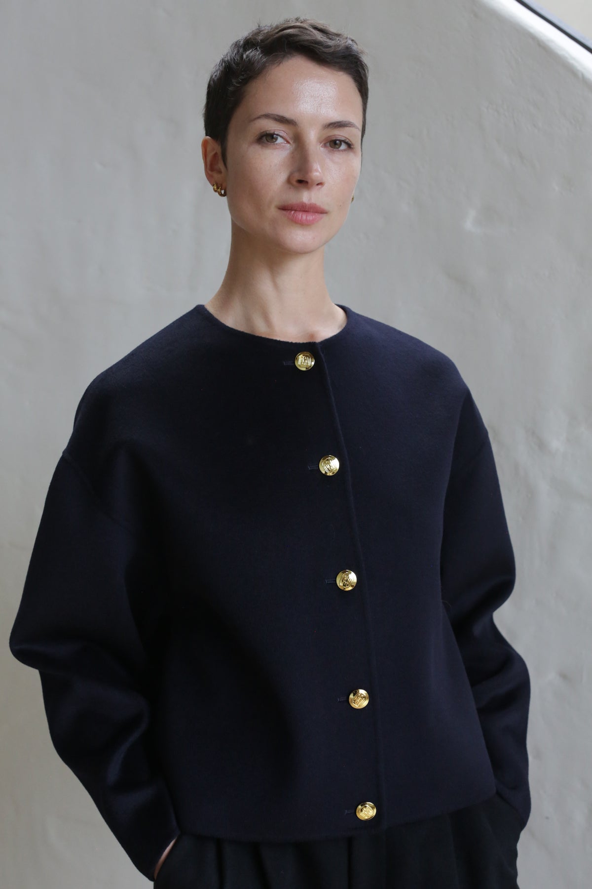 A person with short dark hair and gold stud earrings stands against a light textured wall, wearing the Cocoon Jacket in Navy with gold buttons and hands in pockets, looking calmly at the camera.
