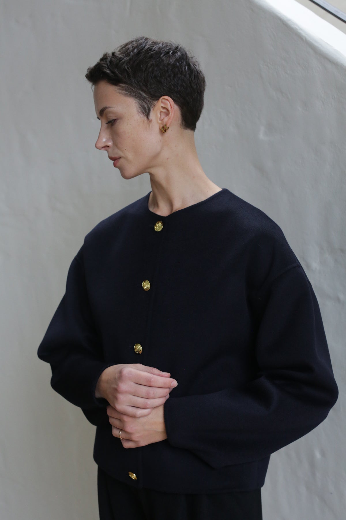 A person with short dark hair stands indoors against a light wall, wearing the Cocoon Jacket in Navy with gold buttons. They look to the side, hands loosely clasped in front.