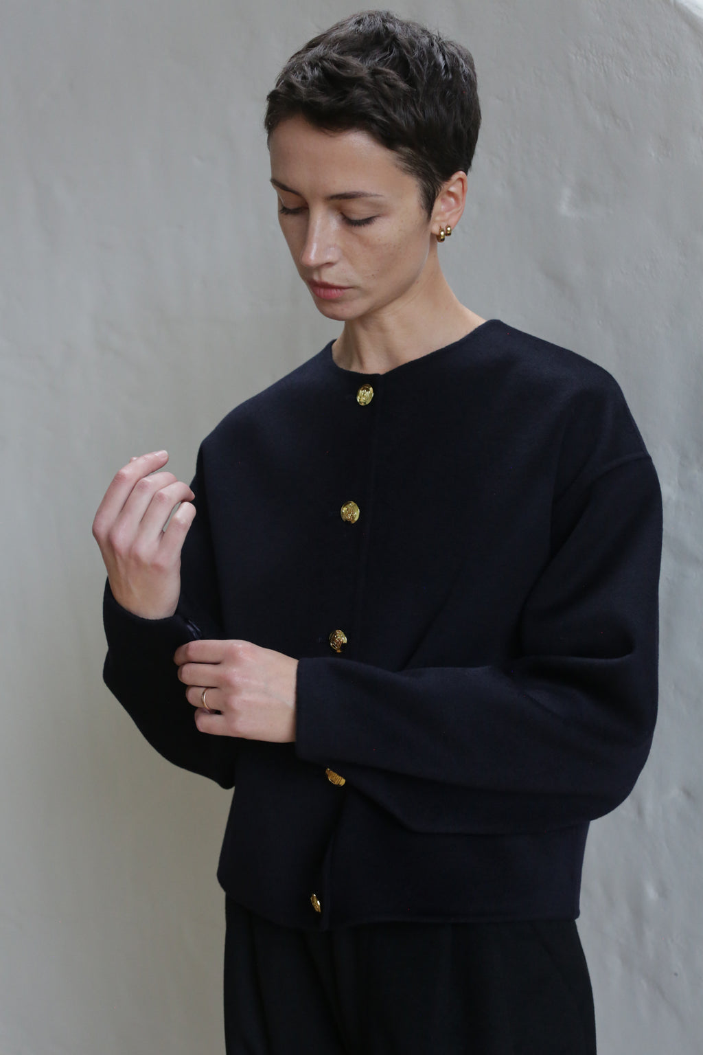 A person with short hair wears the Cocoon Jacket in Navy, standing against a light textured wall, looking down as they adjust their sleeve.