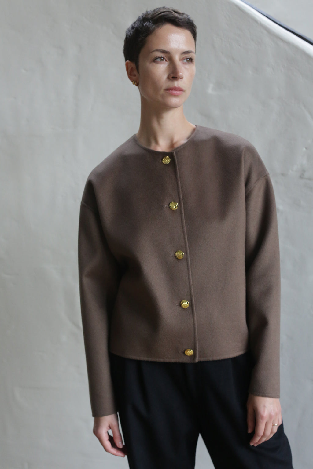 A person with short hair wears the Cocoon Jacket in taupe and black pants, standing before a light-colored wall and gazing slightly to the side.