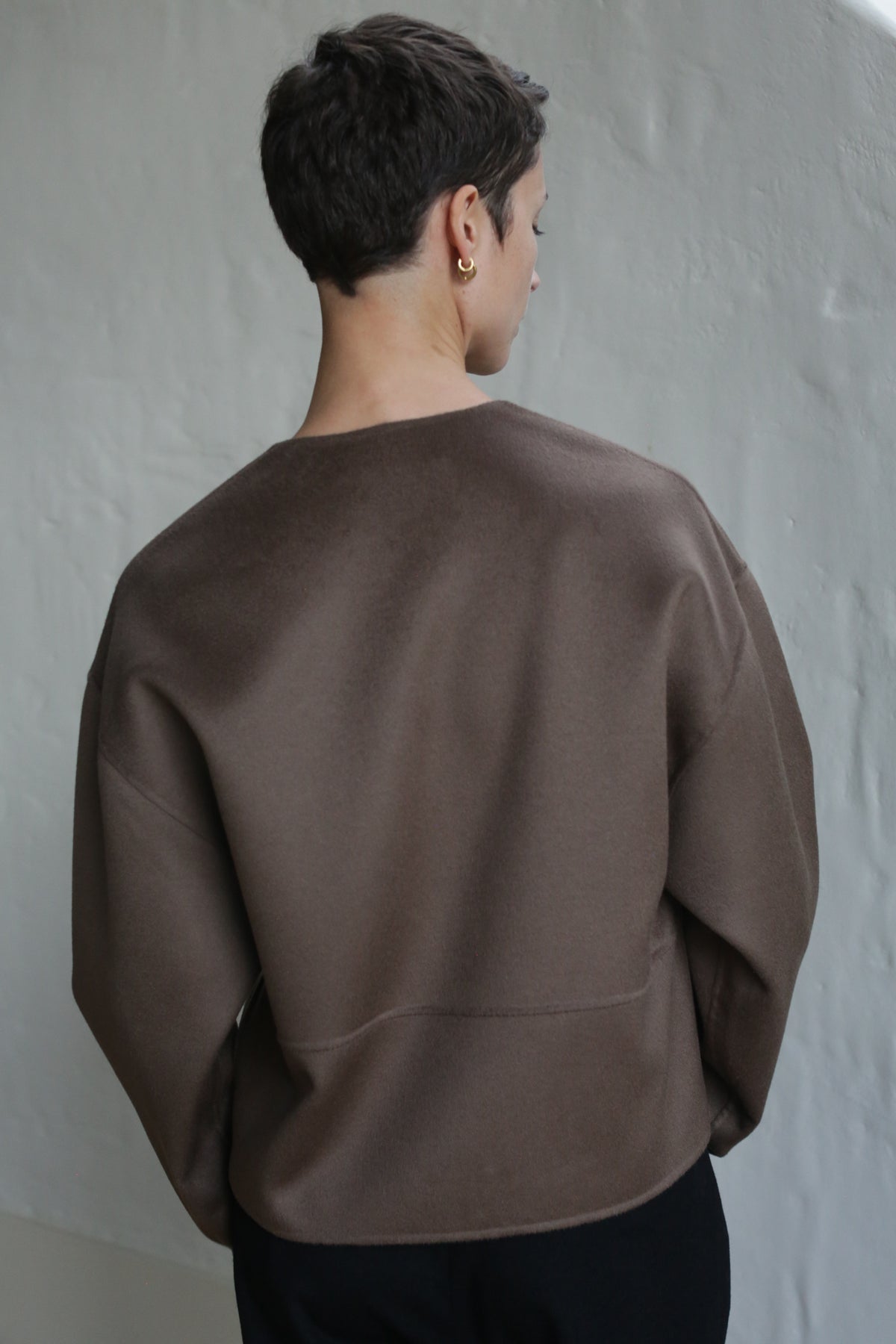 A person with short dark hair, wearing the Cocoon Jacket in Taupe and gold hoop earrings, stands facing away from the camera against a light-colored textured wall.