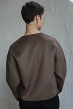 Load image into Gallery viewer, A person with short dark hair, wearing the Cocoon Jacket in Taupe and gold hoop earrings, stands facing away from the camera against a light-colored textured wall.