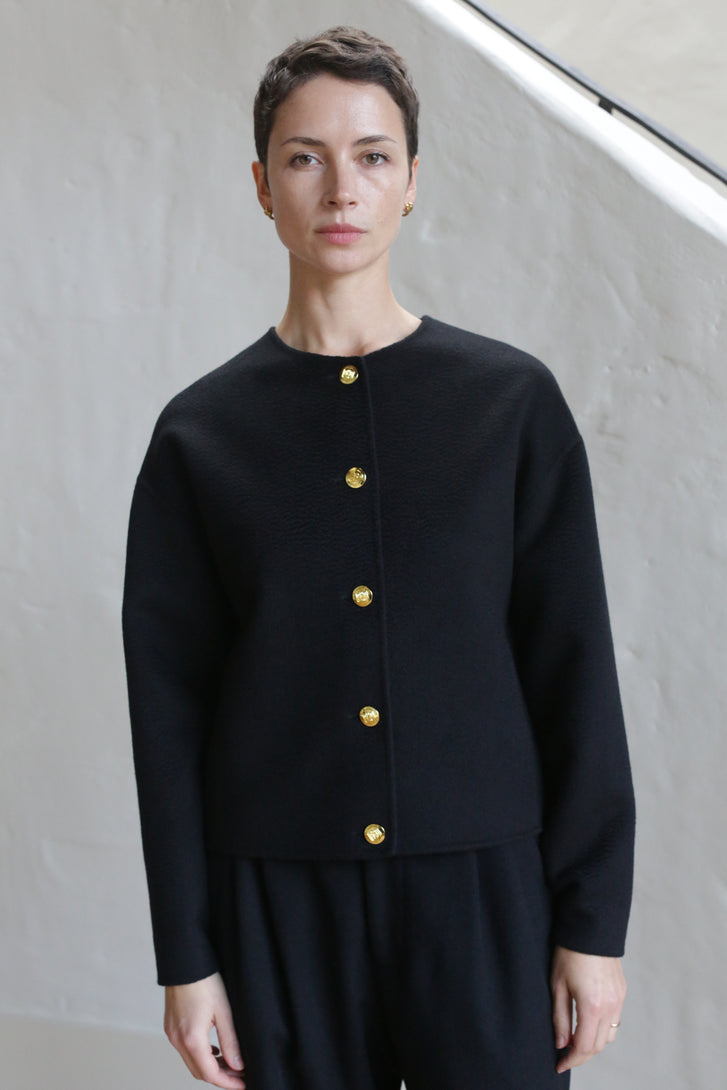 A person with short dark hair wears the Cocoon Jacket | Watermark Black, featuring gold buttons, and stands calmly in front of a light-colored textured wall.