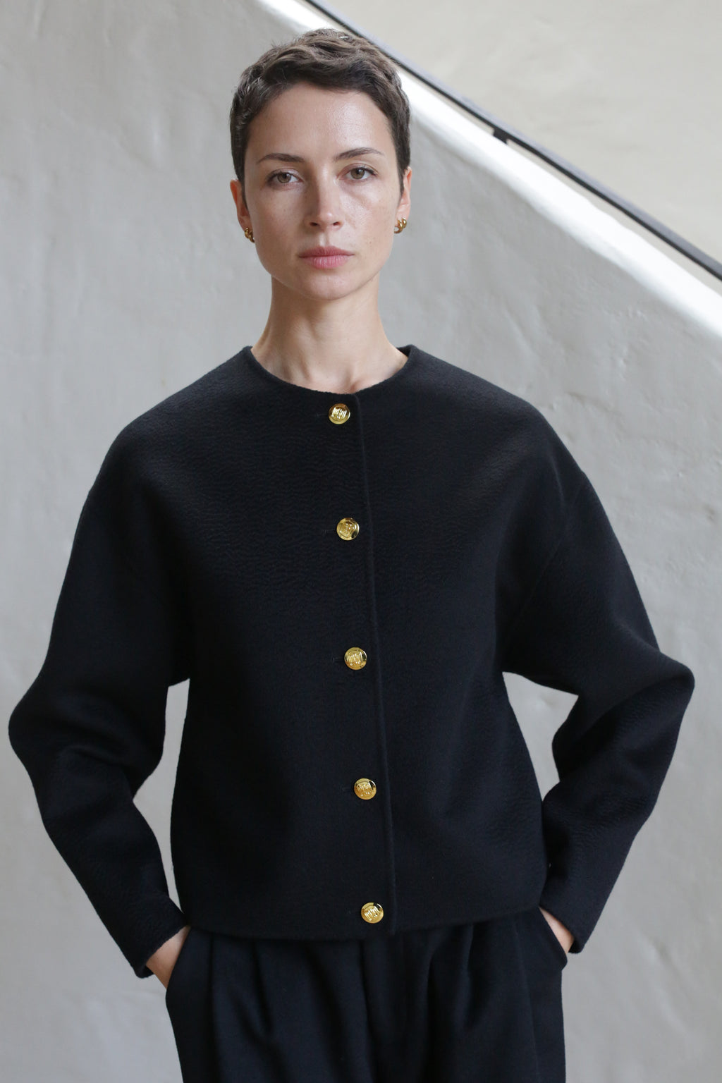 A person with short dark hair stands with hands in pockets, wearing the Cocoon Jacket | Watermark Black, which features gold buttons, against a light-colored wall.