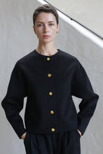 Load image into Gallery viewer, A person with short dark hair stands with hands in pockets, wearing the Cocoon Jacket | Watermark Black, which features gold buttons, against a light-colored wall.