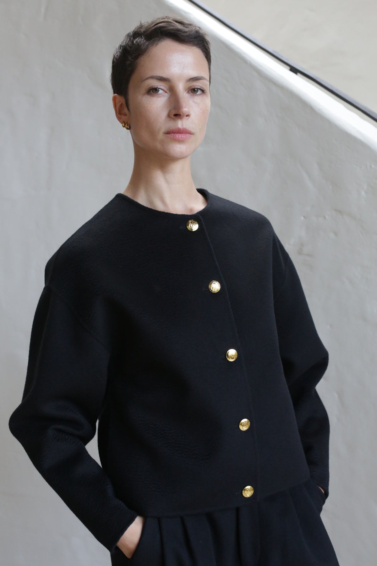 A person with short dark hair wears the Cocoon Jacket | Watermark Black, featuring gold buttons, standing against a light-colored textured wall, looking calmly at the camera with hands in pockets.