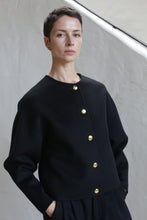 Load image into Gallery viewer, A person with short dark hair wears the Cocoon Jacket | Watermark Black, featuring gold buttons, standing against a light-colored textured wall, looking calmly at the camera with hands in pockets.