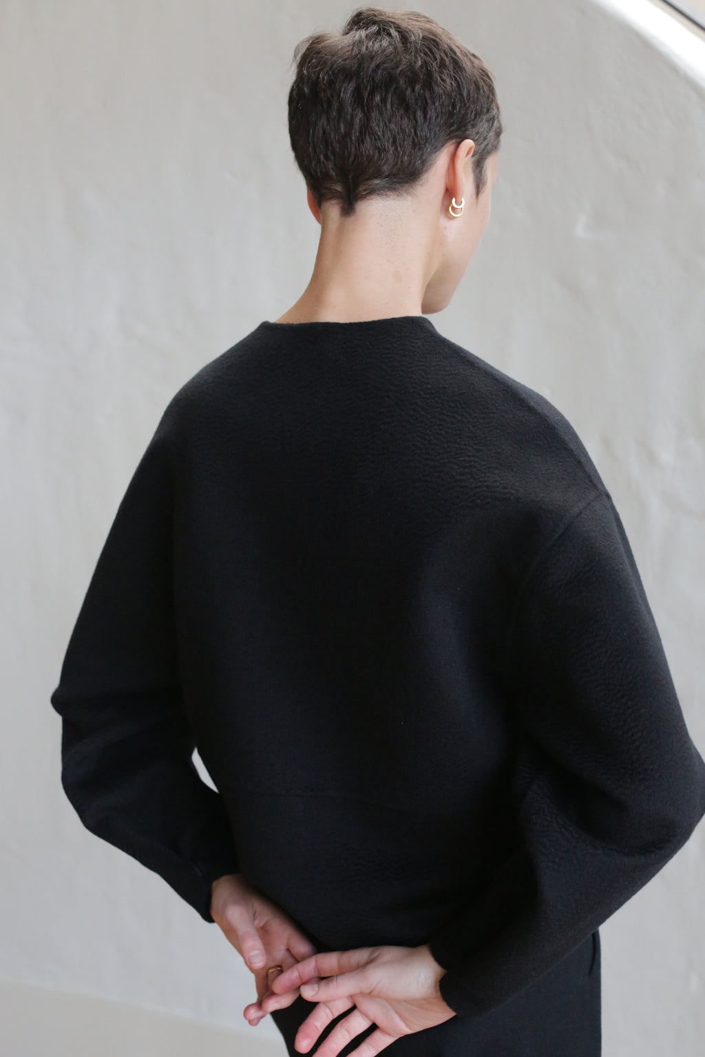A person with short dark hair, wearing the Cocoon Jacket in Watermark Black, stands facing away with hands clasped behind their back against a minimal, light background.
