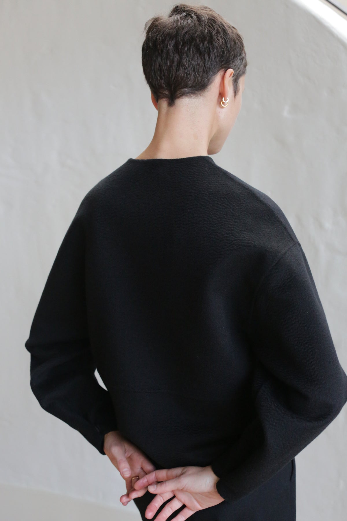A person with short dark hair, wearing the Cocoon Jacket in Watermark Black, stands facing away with hands clasped behind their back against a minimal, light background.