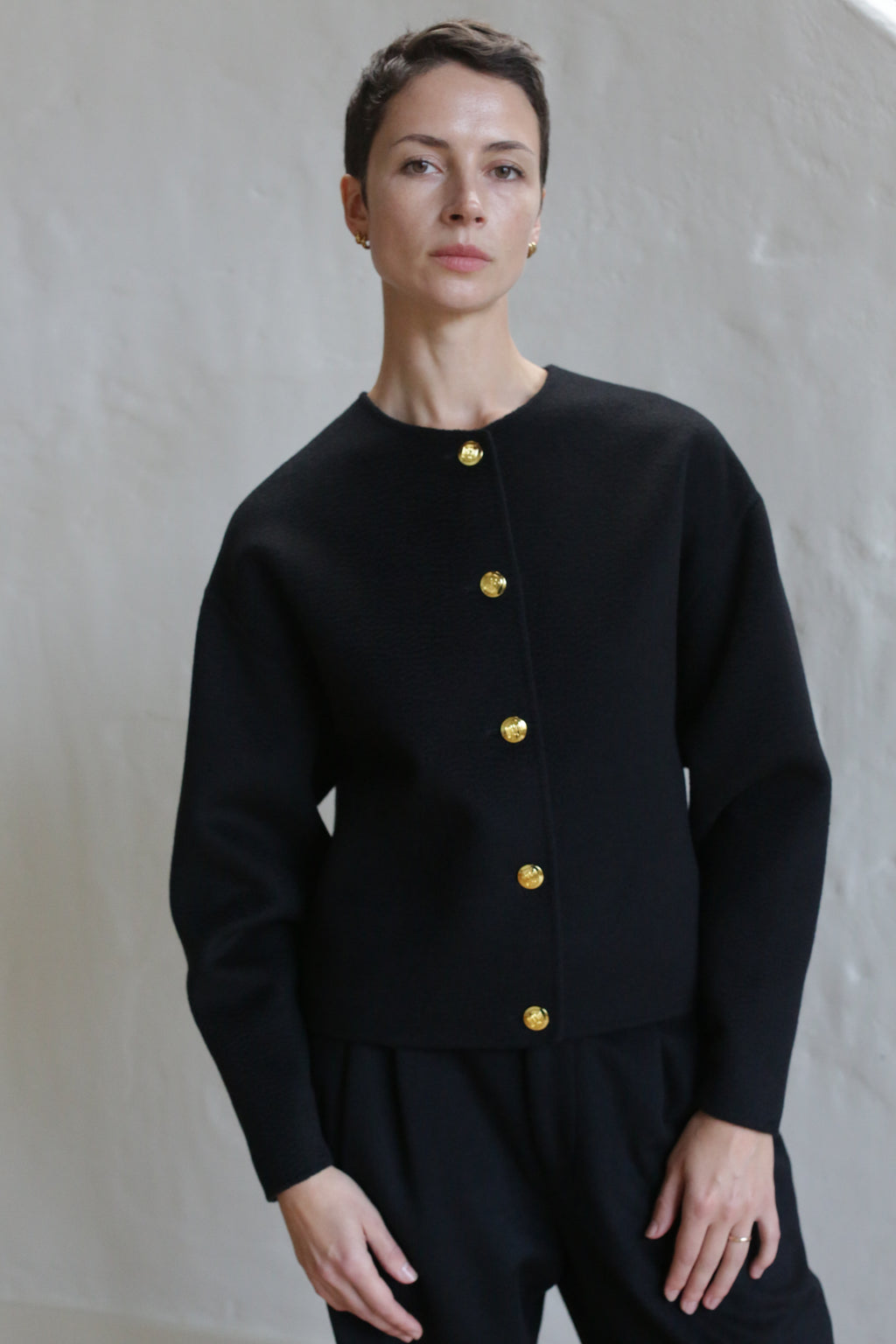 A person with short dark hair wears the Cocoon Jacket in Watermark Black, paired with matching black pants, standing against a light-colored textured wall.