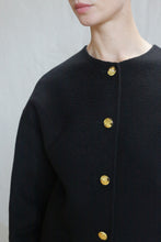 Load image into Gallery viewer, A person wearing the Cocoon Jacket | Watermark Black, featuring a textured black fabric and round gold buttons, shown from shoulders to mid-torso against a plain light background.