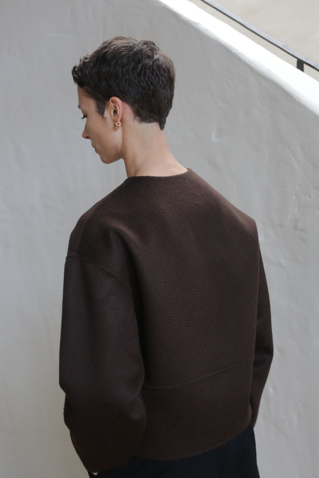 A person with short dark hair, wearing the Cocoon Jacket in Watermark Brown, stands facing away near an off-white wall.