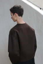 Load image into Gallery viewer, A person with short dark hair, wearing the Cocoon Jacket in Watermark Brown, stands facing away near an off-white wall.