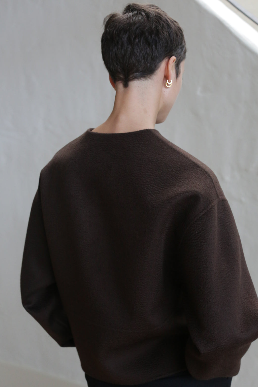 A person with short dark hair models the Cocoon Jacket in Watermark Brown, standing with their back to the camera against a light-colored background.