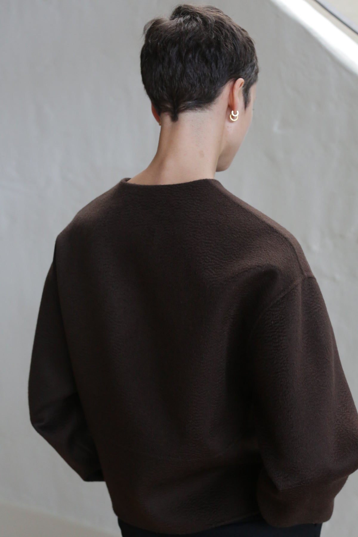 A person with short dark hair models the Cocoon Jacket in Watermark Brown, standing with their back to the camera against a light-colored background.