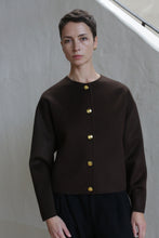 Load image into Gallery viewer, A person with short dark hair wears the Cocoon Jacket in Watermark Brown, featuring long sleeves and five gold buttons, standing against a light-colored wall and looking directly at the camera with a neutral expression.