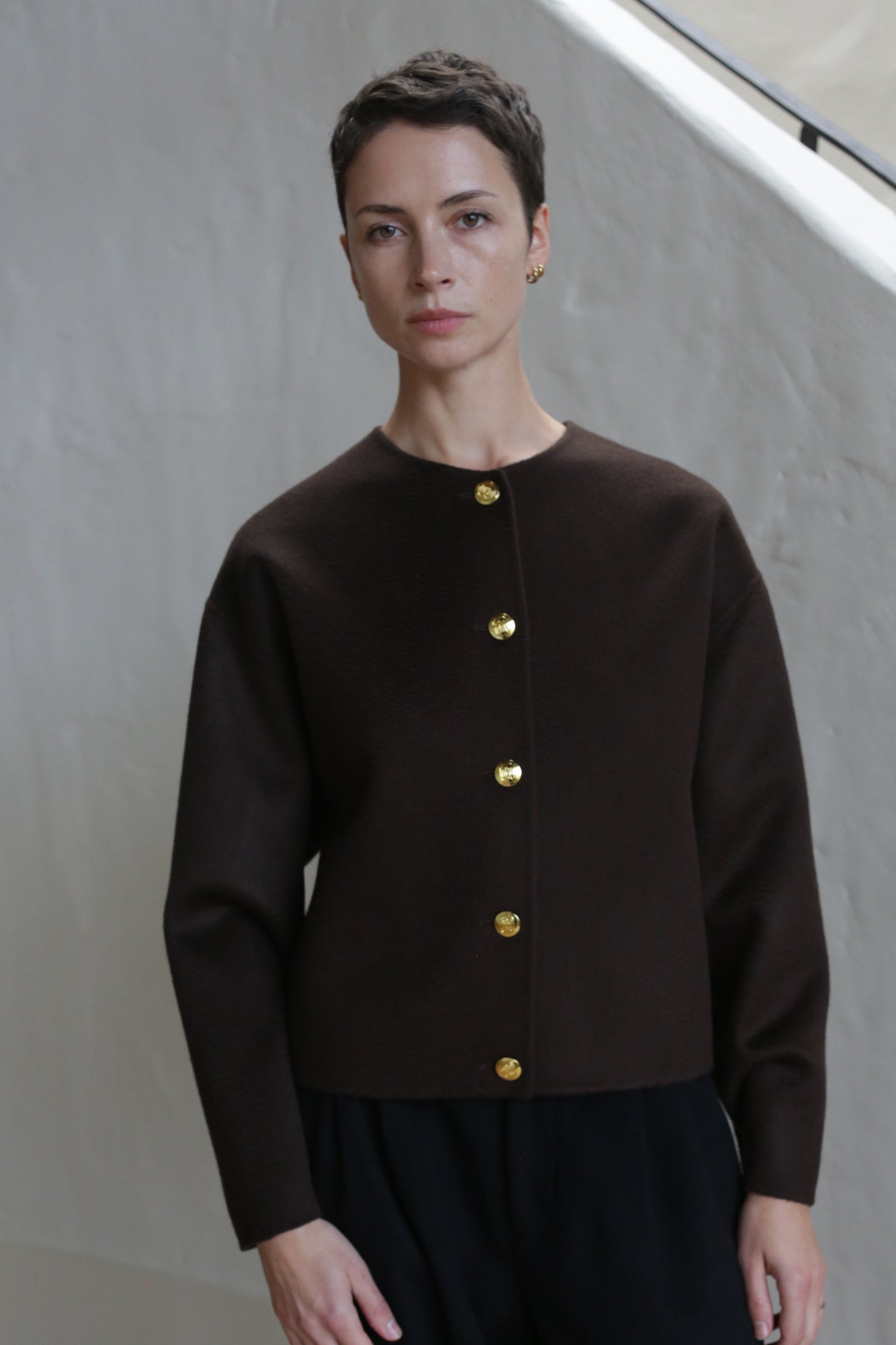A person with short dark hair wears the Cocoon Jacket in Watermark Brown, featuring long sleeves and five gold buttons, standing against a light-colored wall and looking directly at the camera with a neutral expression.