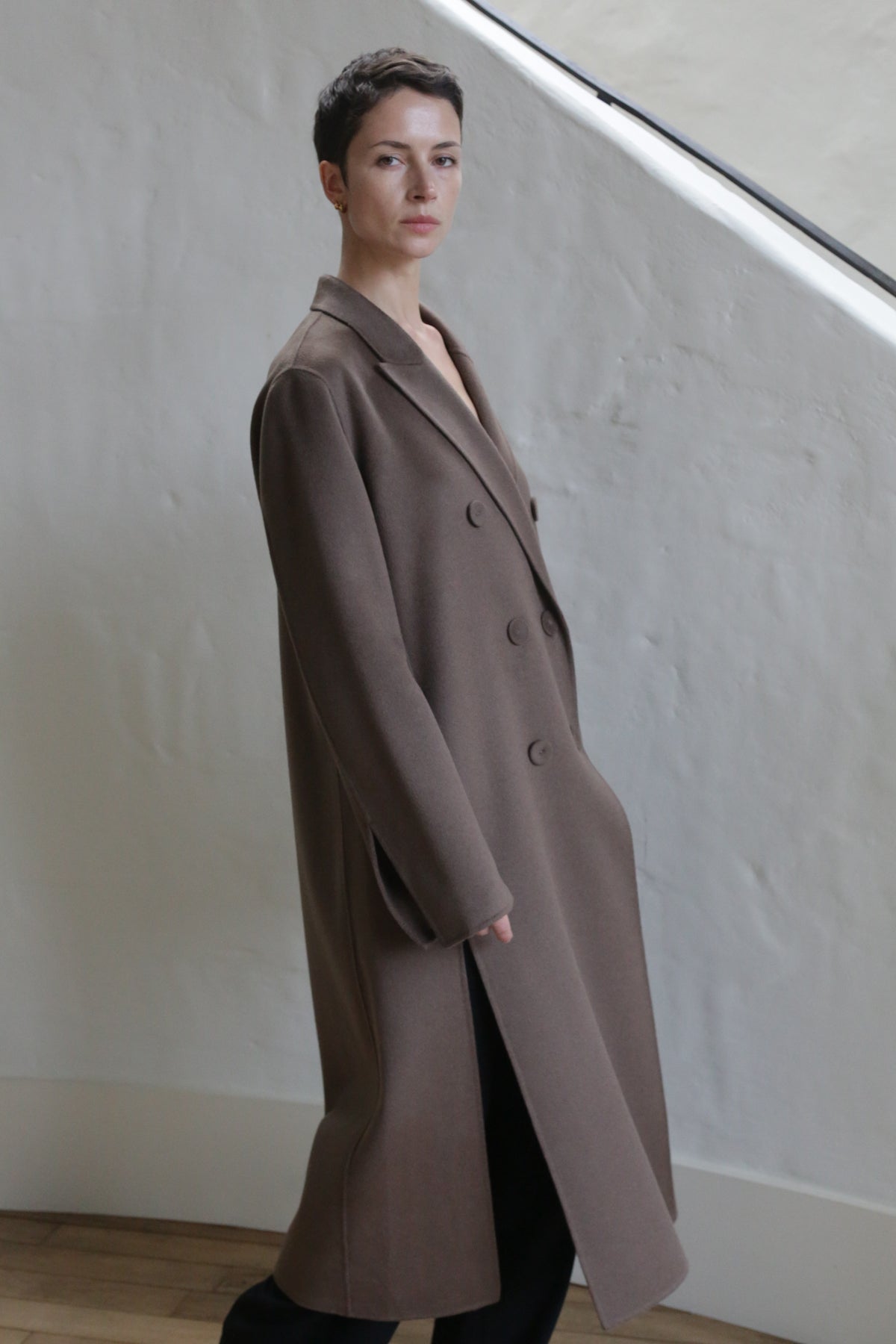 A person with short dark hair wears the Doublebreasted Coat in Taupe and black pants, standing indoors against a white textured wall with wooden flooring and a staircase in the background.