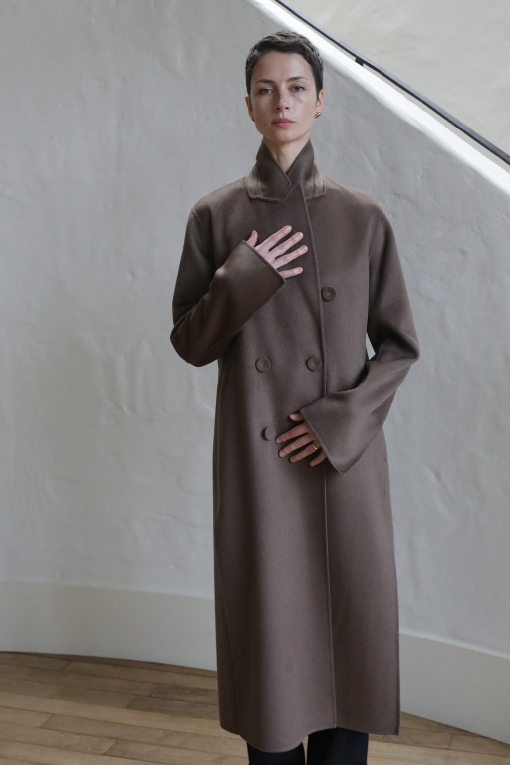 A person wearing the Doublebreasted Coat | Taupe stands indoors against a light textured wall, with one hand on their chest and the other near their waist.