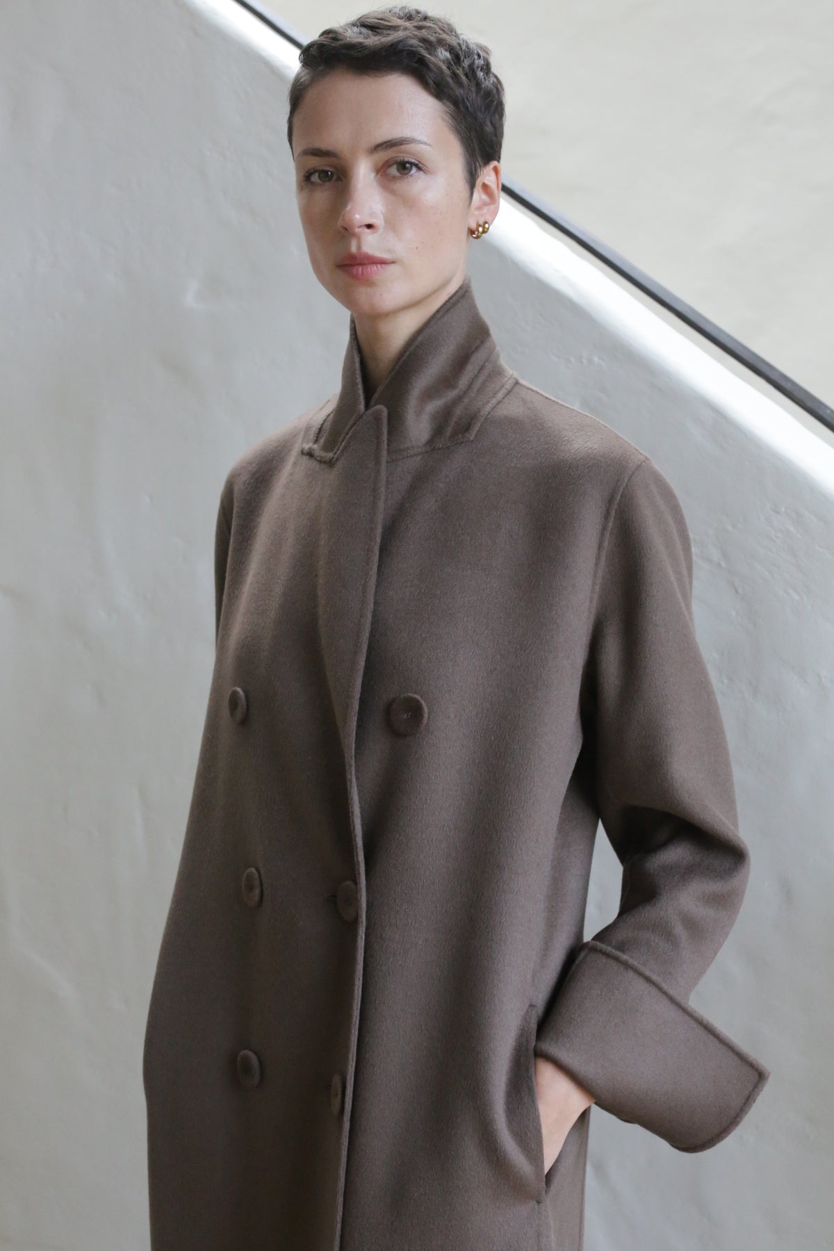 A person with short dark hair, wearing the Doublebreasted Coat in Taupe, stands against a light textured wall with one hand in their pocket, looking calmly at the camera.