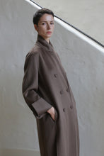 Load image into Gallery viewer, A person with short dark hair wears the Doublebreasted Coat | Taupe and stands with one hand in their pocket against a light-colored textured wall and staircase.
