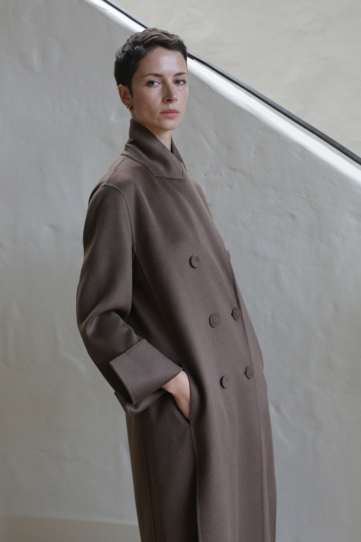A person with short dark hair wears the Doublebreasted Coat | Taupe and stands with one hand in their pocket against a light-colored textured wall and staircase.