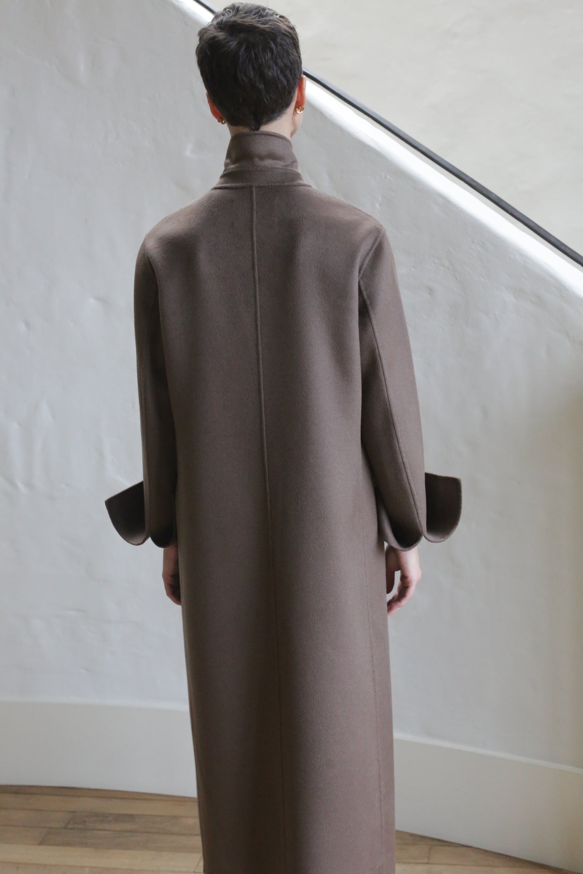 A person with short dark hair stands indoors, facing away, wearing the Doublebreasted Coat | Taupe. The coat features a high collar and wide cuffs. The background shows a white textured wall and wooden floor.